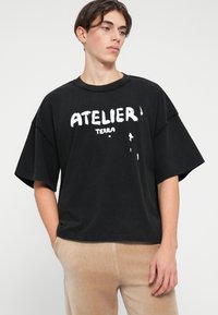 Črna oversize majica s kratkimi rokavi, z belim napisom "ATELIER TERRA" v postarane različici. Mehka bombažna tkanina.