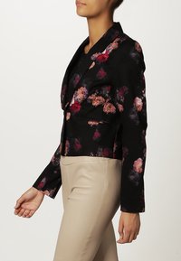 Femme portant un blazer floral noir avec des fleurs rouges et roses, associé à un pantalon beige taille haute, posant de profil devant un fond uni.