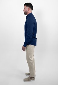 Marineblauwe blouse met lange mouwen en een button-down kraag, gecombineerd met crèmekleurige chino's en beige instappers. Witte achtergrond.