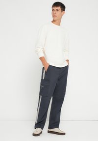 Balta garām piedurknēm krekls kopā ar tumši ziliem cargo biksēm ar sānu strīpām un vairākiem kabatām, papildināts ar krēmkrāsas sporta kurpēm.