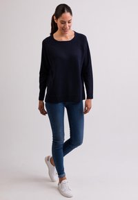 Pull en tricot bleu marine avec un col large, associé à un jean bleu ajusté et des baskets blanches, présentant un design épuré et une coupe décontractée.