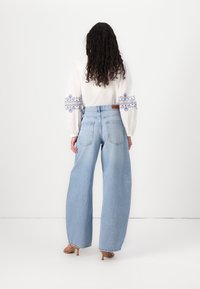Jeans larghi di colore blu chiaro, in cotone, a vita alta, abbinati a una camicetta bianca con ricami floreali blu e maniche a sbuffo.