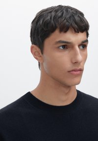 Schwarzer Rundhalspullover aus weichem Material, strukturiertes Strickmuster, taillierte Passform. Das Modell hat kurze Haare und einen neutralen Gesichtsausdruck.