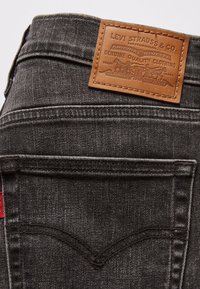 Jeans in denim nero con superficie testurizzata. Presentano una toppa in pelle beige con logo in rilievo, tasche posteriori e un'etichetta rossa del marchio.
