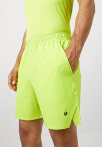 Hombre que lleva pantalones cortos deportivos de un brillante color verde lima, con bolsillos laterales, cintura elástica y un pequeño logo negro en la pierna izquierda.