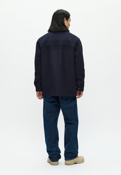 Navy blue jacket made of sturdy fabric, featuring a slightly oversized design, buttoned cuffs, and a smooth texture. Worn with blue denim pants.