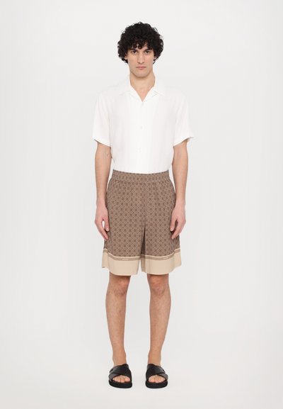 Camicia bianca a colletto con maniche corte abbinata a pantaloni corti marroni con motivo e orlo beige uniforme. Il modello indossa sandali neri.