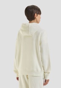 Felpa color crema con una texture morbida, dotata di cappuccio regolabile con cordino e polsini a coste. Vestibilità rilassata con design a spalla scesa.