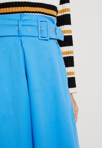 En blå kjol med en bred midja med ett rektangulärt bälte och silverdetaljer, tillverkad av ett mjukt tyg med en ren, minimalistisk design.