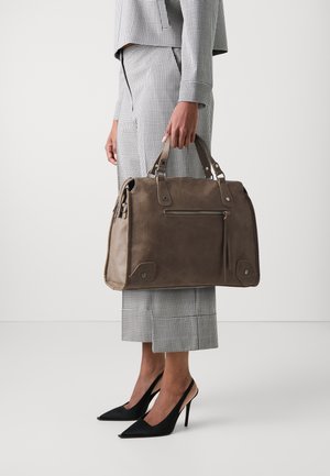 Persona con un traje de cuadros en blanco y negro y tacones altos negros, sosteniendo un gran bolso de cuero marrón con bolsillo con cremallera.