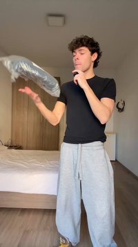 Νέος άντρας με μαύρο πουκάμισο και γκρί φόρμα πετάει μια κλειστή πλαστική συσκευασία σε ένα υπνοδωμάτιο με ξύλινα έπιπλα.