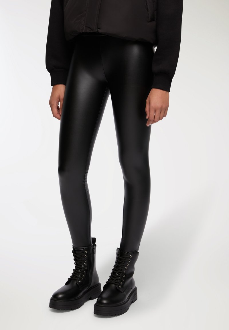 Leggings in finta pelle nera con una finitura liscia e lucida. Abbinati a stivali combat neri con lacci, caratterizzati da suole spesse e occhielli metallici.