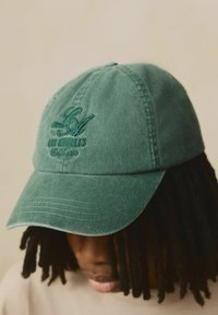 Gorra de algodón verde con bordado de "LA" y "Los Ángeles, California." Presenta visera curvada y tejido texturizado.
