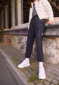Black tapered cargo pants with elastic cuffs, featuring side pockets. Worn with a white quilted jacket and white sneakers on a textured surface.