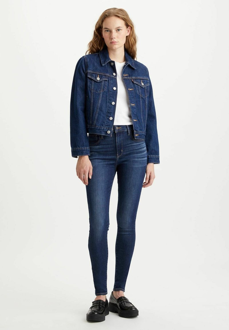 Chaqueta de mezclilla azul oscura con un corte clásico, con cierres de botón y dos bolsillos frontales, combinada con pantalones skinny azules y zapatos de plataforma negros.