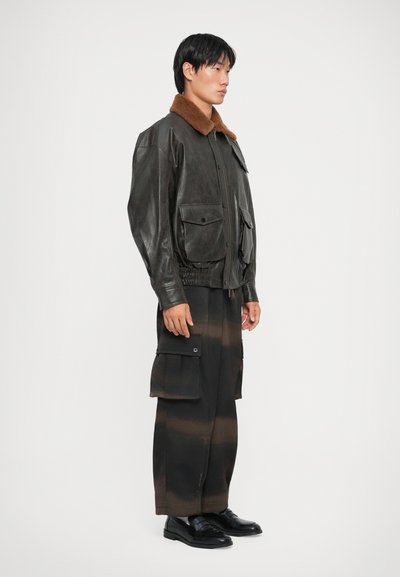 Brown leather jacket with a faux fur collar, featuring two front pockets and an elastic waistband, paired with dark, striped cargo pants.