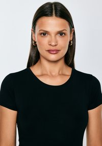 Jonge vrouw met stijlbruin haar, goudkleurige oorringen en een zwart shirt met korte mouwen, die recht vooruit kijkt tegen een effen achtergrond.