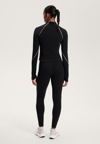 Fekete, hosszú ujjú sportfelső, fényvisszaverő részletekkel, fekete leggings-szel párosítva. Az anyaga rugalmas, testhezálló, ujjlyukakkal.