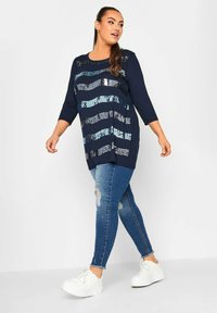 Tunika navy con strisce orizzontali di paillettes, maniche a trequarti, abbinata a jeans strappati e sneaker bianche. Tessuto morbido.