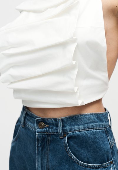 Λευκό ασύμμετρο τοπ με μεγάλο βολάν, φορεμένο με ψηλόμεσο μπλε τζιν, που δείχνει την κοιλιά και το πλάι του κορμού.