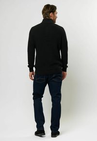 Črn, rebrast pulover z visokim ovratnikom, v kombinaciji z temno denim hlačami. Manekenka prikazana od zadaj, kar poudarja teksturo in prileganje puloverja.