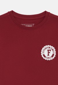 Camiseta de algodón color marrón con un escote redondo, que presenta un logo circular blanco con "MANHATTAN NEW YORK COLLEGE" y una "F" prominente.