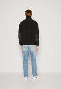 Giacca in pile nera con colletto alto e maniche scure, abbinata a jeans azzurri chiari e sneakers bianche. Design semplice, senza motivi visibili.