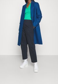 Blauwe wollen blend jas over een groene geribbelde top, gecombineerd met donkere broeken en witte platform sneakers. Over het geheel een vlakke, gladde textuur.