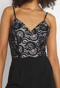 Svart kamisol med spetsöverdrag som har en v-ringning och tunna axelband, prydd med intrikata blommiga och spiralformade mönster.