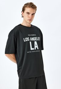 Camiseta negra de manga corta hecha de tela suave, con texto blanco: "CALIFORNIA LOS ÁNGELES LA ESTADOS UNIDOS ATLETISMO."