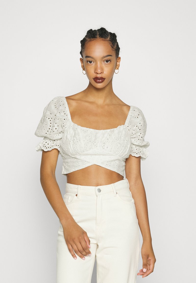 Blusa branca curta com mangas bufantes e decote quadrado, apresentando bordado de olhal. Combinada com calças de cintura alta de cor clara.