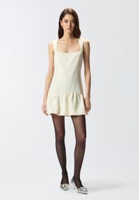 Vestido mini blanco con escote cuadrado, dobladillo con volantes y corsé ajustado. La modelo lleva medias negras translúcidas y tacones plateados adornados.