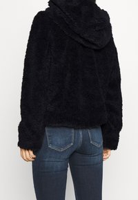 Svart, fluffig fleecejacka med huva, kort design och långa ärmar, som är kombinerad med mid-rise blå jeans med bakfickor.