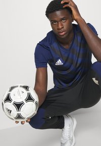 Dunkelblau gestreiftes Adidas-Trikot, schwarze Hose, hält einen schwarz-weißen Fußball. Mit Logo und modernem sportlichen Schnitt.