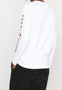 T-shirt bianco a maniche lunghe con ricamo del testo "TOMMY HILFIGER" sulla manica sinistra. Realizzato in morbido tessuto di cotone. Design semplice e pulito.