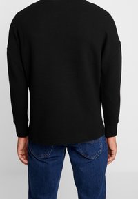 Maglietta nera a maniche lunghe con texture a costine, vestibilità rilassata, orlo arrotondato e maniche con polsini, abbinata a jeans in denim blu.
