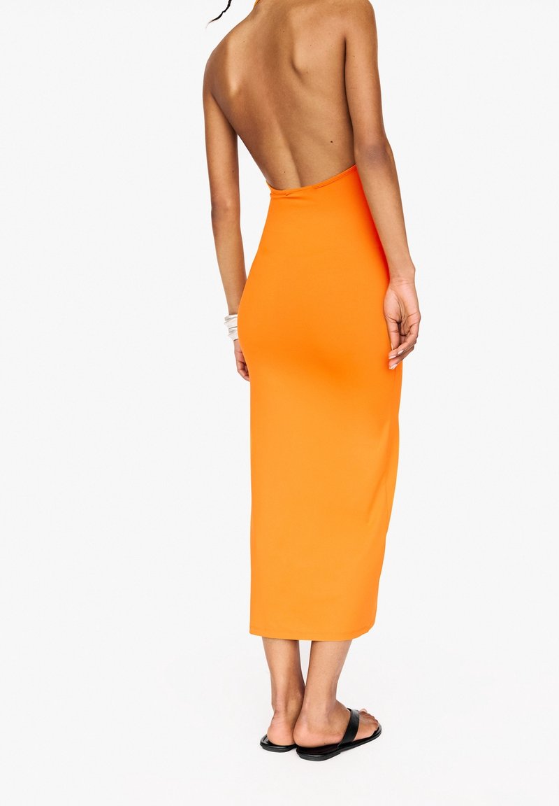 Femme portant une robe midi orange dos nu et sans manches, avec des sandales plates noires, debout sur un fond blanc uni.