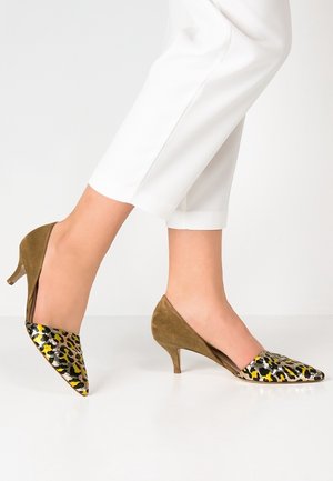 Voeten in bruine suède kitten heels met geel-zwarte luipaardprint op de puntige neuzen, gecombineerd met witte cropped broeken.