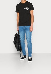 Černé tričko s bílým logem "Calvin Klein Jeans", zkombinované s modrými úzkými džíny a černými teniskami, držící černý batoh.