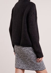 Schwarzer, grob gestrickter Pullover mit hohem Kragen, kombiniert mit einem gemusterten Rock in Schwarz und Weiß im Hahnentrittmuster, der strukturierten Stoff zeigt.