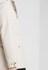 Vue latérale d'une personne portant une veste beige à capuche avec une fermeture éclair à l'avant et des poches à rabat à boutons-pression.