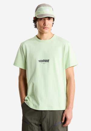 Mand iført en lysegrøn t-shirt med "The North Face" logo og en matchende cap, stående med den ene hånd i lommen mod en hvid baggrund.