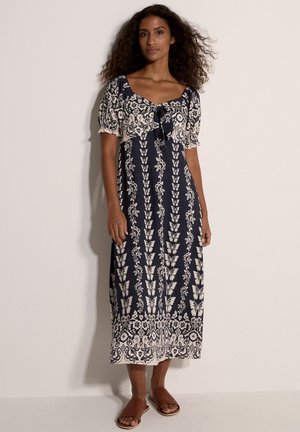 Femme aux cheveux bouclés portant une robe midi bleu marine et crème à motifs floraux et papillons, ainsi que des sandales mules marron, debout contre un mur uni.