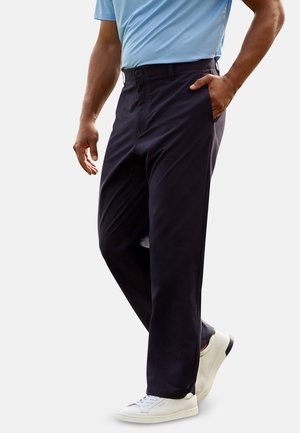 Hombre vistiendo pantalones azul marino oscuro con bolsillos laterales, camisa azul claro y zapatillas blancas, de pie con una mano en el bolsillo.