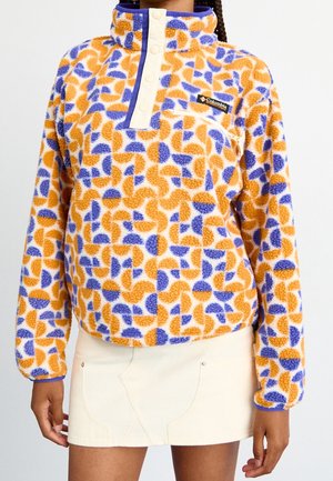 Mujer que lleva un pulóver de forro polar con un patrón abstracto en naranja y azul, abertura con botones a presión y minifalda blanca.