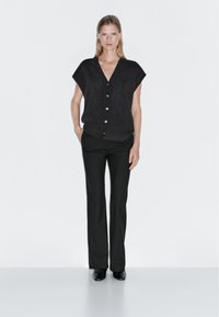 Frau in einem dunklen, kurzärmeligen Knopf-Vest, schwarzen geraden Hosen und schwarzen Schuhen, die vor einem schlichten, hellen Hintergrund steht.