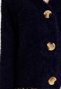 Navy blue, fuzzy knit cardigan with three large, swirl-patterned beige buttons. Soft texture, deep V-neck, and chunky knit design.