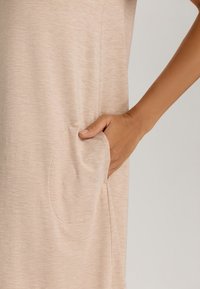Abito beige con un tessuto morbido e lavorato. Presenta una tasca arrotondata sul lato, con una mano all'interno della tasca. Design semplice.