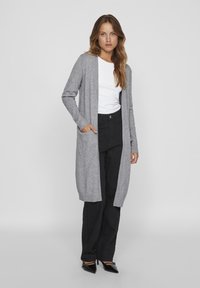 Cardigan long gris avec des poignets côtelés et une ouverture sur le devant, porté sur un t-shirt blanc et un pantalon noir, comportant des poches latérales.