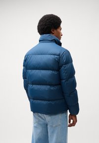 Jachetă puffer de un albastru închis, cu un guler înalt, design matlasat și textură netedă, purtată cu blugi din denim de un albastru deschis.